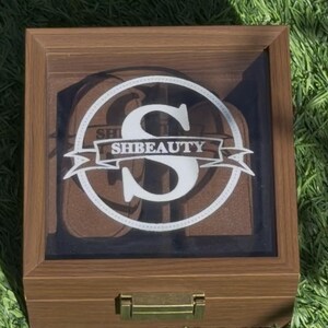 May include: A wooden box with a clear lid, displaying a white circular design with the letter "S" and the text "SHBEAUTY". The box has a brass-coloured latch and is set against green grass.
