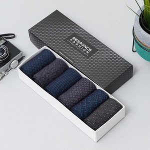 May include: A set of six patterned socks in a gift box. The socks are in shades of blue and grey, with various designs. The box has a black, textured lid with the text "RESPONCE FASHION ACCESSORIES FOR MEN". A camera and a plant are visible.