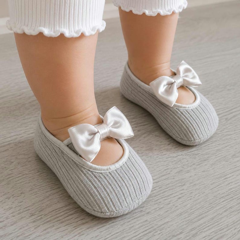 A infant wearing white pants and gray ballerina socks with bows stands on a light wooden floor.