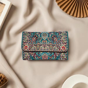 May include: A teal wallet with a detailed, colourful floral pattern. The wallet has a fold-over flap and appears to be made of a woven fabric. The design incorporates red, white, and purple accents, creating a vintage aesthetic.