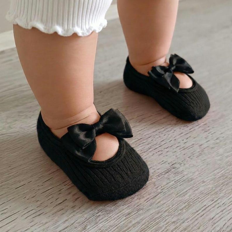 An infant wearing white pants and black socks with bows stands on a light wooden floor.