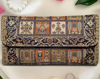 Op de afbeelding: Een rechthoekige clutch met een gedetailleerd patroon in marineblauw, oranje en crème. De clutch heeft een overslagklep en een goudkleurige rand. Het ontwerp bevat bloemen en geometrische motieven.