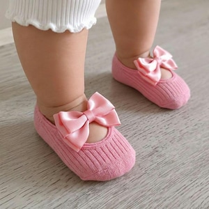 A toddler wearing white pants and pink socks with bows stands on a light wooden floor.