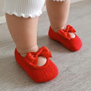 A baby wearing red baby girl socks with bows stands on a light wooden floor.