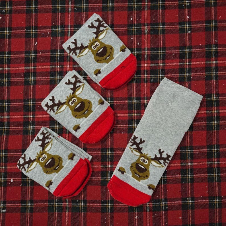 May include: Four gray socks with a reindeer design and red toes are arranged on a red and black plaid surface. The reindeer has brown antlers and a smiling face. The socks are likely for the holiday season.