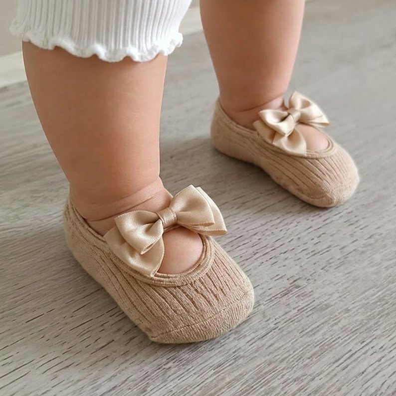 A baby wearing beige ballerina socks with bows stands on a light wooden floor.
