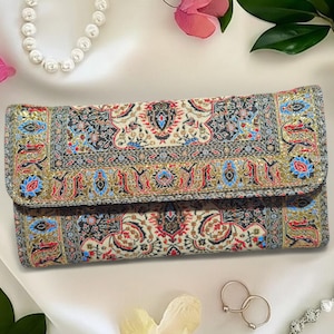 May include: A rectangular clutch with a detailed, colorful pattern. The design features red, blue, black, and gold accents on a cream background. The clutch has a fold-over flap and is likely made of fabric.