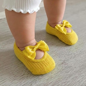 A baby wearing yellow socks with bows stands on a light wooden floor.