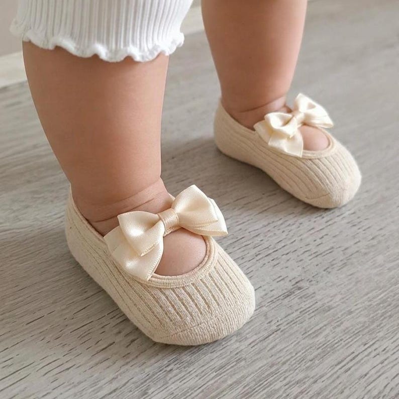A baby wearing cream white socks with bows stands on a light wooden floor.