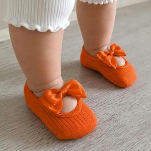 A toddler wearing white pants and orange socks with bows stands on a light wooden floor.