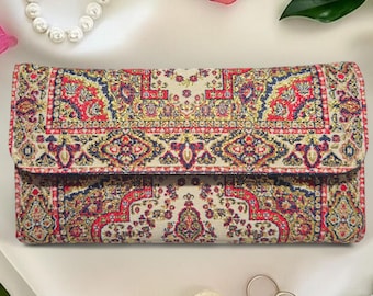 Op de afbeelding: Een rechthoekige clutch met een kleurrijk, ingewikkeld patroon in rood, blauw, geel en crème. Het ontwerp lijkt op een traditioneel tapijt of textiel. De clutch sluit met een flap en lijkt van stof te zijn.
