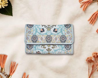 May include: A light blue wallet with a floral pattern in shades of blue, pink, and gold. The wallet has a rectangular shape with a fold-over flap. The design features symmetrical floral and geometric elements.
