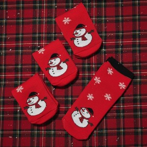 May include: Four red Christmas socks with a snowman design and white snowflakes. The snowman has a black hat, red scarf, and black button eyes. The socks are on a red plaid background.
