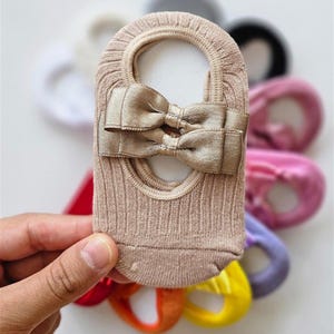 Op de afbeelding: Een beige onzichtbare sok met een decoratieve strik. De sok heeft een geribbelde textuur en een kleine opening aan de bovenkant. Andere sokken in verschillende kleuren zijn zichtbaar op de achtergrond.