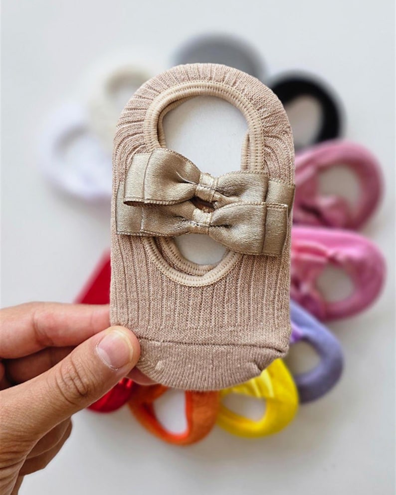 Op de afbeelding: Een beige onzichtbare sok met een decoratieve strik. De sok heeft een geribbelde textuur en een kleine opening aan de bovenkant. Andere sokken in verschillende kleuren zijn zichtbaar op de achtergrond.