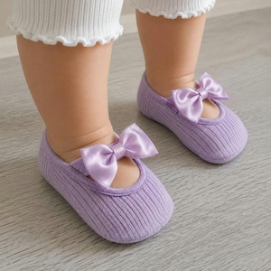 An infant wearing lilac ballerina socks with bows stands on a light wooden floor.