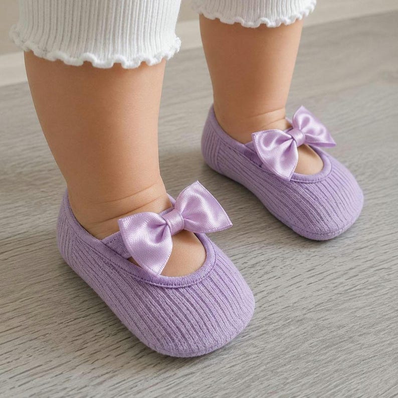 An infant wearing lilac ballerina socks with bows stands on a light wooden floor.