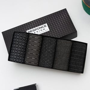 以下が含まれることがあります： 黒いギフトボックス、テクスチャード加工の蓋付き。5足の柄入り靴下が入っています。靴下は黒とグレーの色合いで様々なデザインが施されています。「RESPONCE DESIGN」と「DIES FOR MEN」の文字が入っています。ヴィンテージカメラが手前にあります。