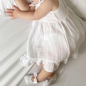 A baby girl wearing a white dress and ballerina socks with bows is sleeping on a white bed, lying on their side with one arm bent and head resting on a pillow.