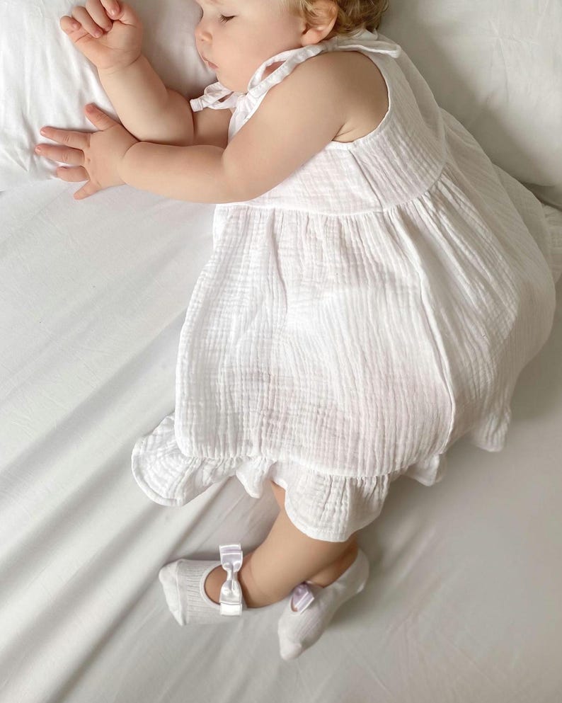 A baby girl wearing a white dress and ballerina socks with bows is sleeping on a white bed, lying on their side with one arm bent and head resting on a pillow.