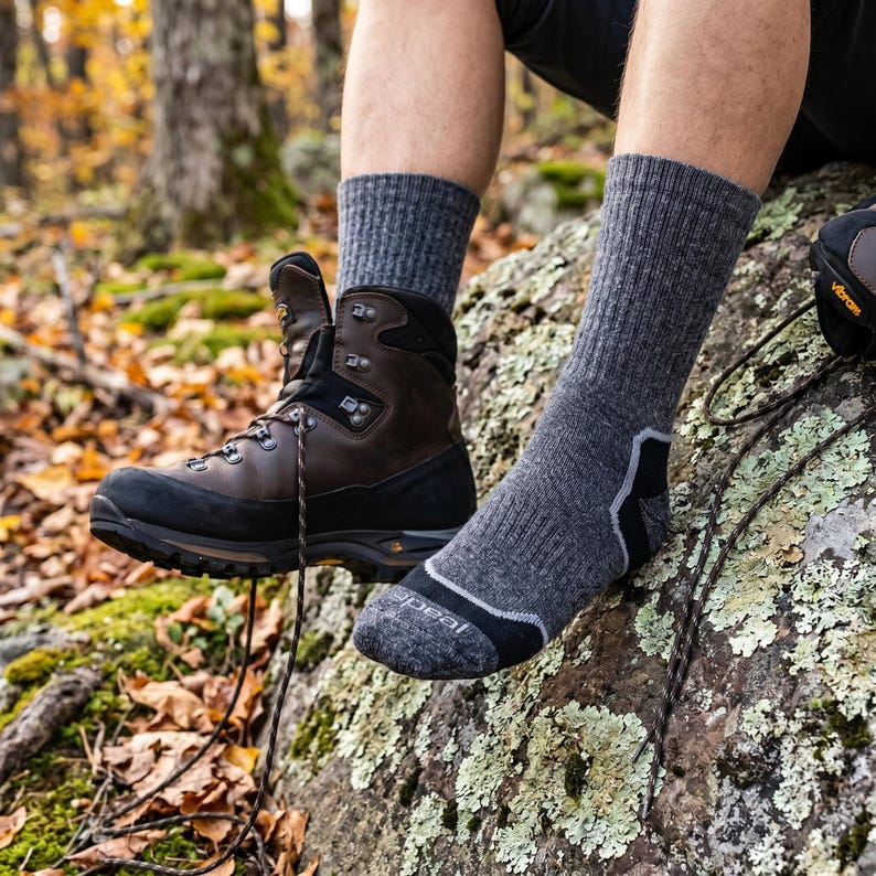May include: A pair of mid-calf charcoal gray wool socks with black reinforced toe and heel sections, featuring the word 'peal' printed on the toe. One foot is bare, while the other wears a brown leather hiking boot with black rubber trim.