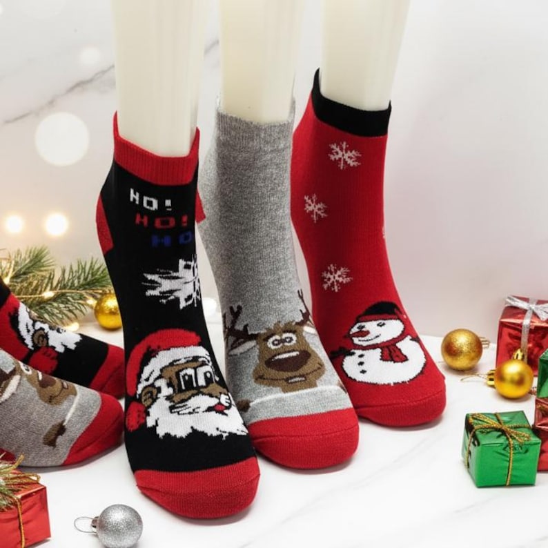 Colorful Christmas-themed ankle socks featuring Santa Claus, reindeer, snowman, and snowflake designs are displayed on mannequin feet, surrounded by festive holiday decorations and gift boxes.