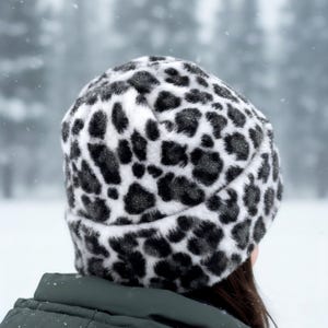 May include: A black and white leopard print faux fur hat. The hat is viewed from the back, set against a blurred, snowy backdrop. A winter accessory.