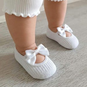A newborn wearing white pants and white socks with bows stands on a light wooden floor.