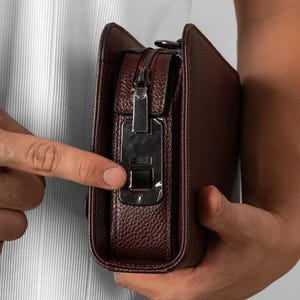 May include: A dark burgundy textured leather clutch bag featuring a rectangular silver-toned metal clasp mechanism on the side. The bag has a zippered closure along the top edge and a structured, rounded rectangular profile with visible stitching.
