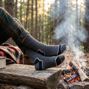 May include: A pair of charcoal gray crew socks featuring a ribbed texture, black reinforced toe and heel sections, and light gray accent lines. The socks are worn with green and brown plaid trousers and a red and black plaid blanket.