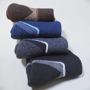 May include: A stack of four pairs of folded knit socks in varying dark colors. The top pair is brown, followed by a blue pair, a charcoal gray pair, and a black pair at the bottom. Each sock features a distinct geometric accent stripe.