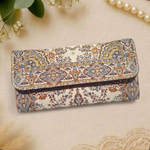 Puede incluir: Una cartera rectangular con un patrón colorido e intrincado que recuerda a una alfombra tradicional. El diseño presenta un fondo crema con motivos florales y geométricos en tonos azules, naranjas y amarillos. La cartera tiene una solapa.