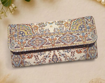 May include: A rectangular wallet with a colorful, intricate pattern reminiscent of a traditional rug. The design features a cream background with floral and geometric motifs in shades of blue, orange, and yellow. The wallet has a fold-over flap.