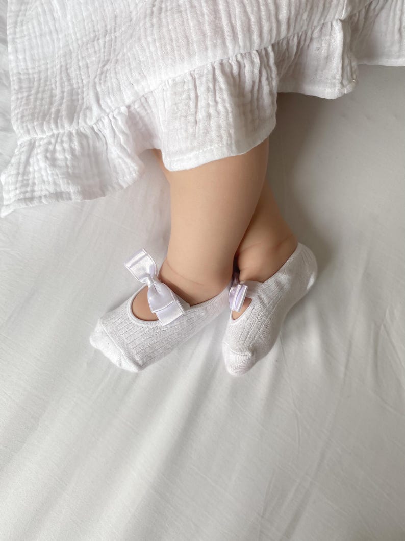 A baby girl wearing white mary-jane style socks with small bows lays on a white surface, her tiny legs are crossed in her sleep.