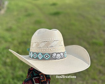 Könnte beinhalten: Ein weißer Strohhut im Cowboy-Stil mit einem türkisfarbenen und weißen Perlenband. Das Band zeigt ein geometrisches Muster und ein Herzdesign. Der Hut wird von einer Hand gehalten, die einen schwarzen und roten gemusterten Ärmel trägt.