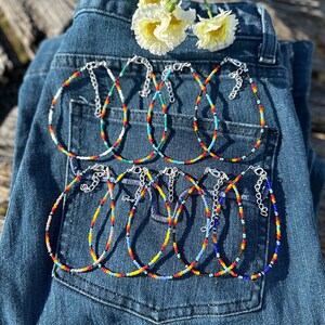 May include: A set of eight colorful beaded anklets with silver clasps. The anklets are arranged on a pair of blue denim jeans.