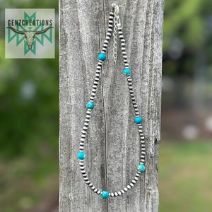 May include: A silver chain necklace with turquoise beads. The necklace is hanging on a wooden post.