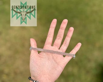 Puede incluir: Primer plano de una mano sosteniendo una pulsera a cuadros rojos y blancos. La pulsera está hecha de pequeñas cuentas cuadradas y tiene un cierre plateado.