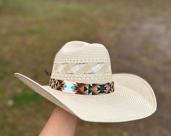 Pode incluir: Um chapéu de cowboy de palha branco com uma faixa de contas com padrões geométricos turquesa, marrom e branco.