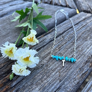 May include: A silver chain necklace with a small silver cross charm and turquoise stones. The necklace is lying on a wooden surface with white flowers.