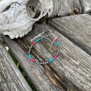 Puede incluir: Un brazalete de cuentas negras y blancas con detalles turquesa y rosa. El brazalete está sobre una superficie de madera.