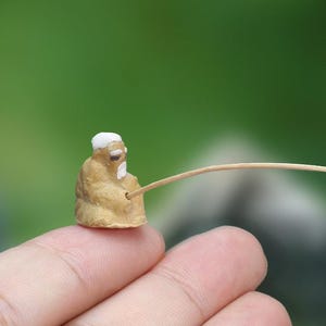 Pode incluir: Uma estatueta em miniatura de cor castanha clara de uma pessoa idosa com cabelo e barba brancos, vestindo uma túnica castanha clara. A figura segura um pau de madeira fino e curvo que se estende para a direita, assemelhando-se a uma cana.