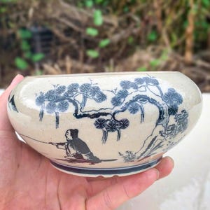 Può includere: Una ciotola in ceramica color crema con un design di ispirazione asiatica blu e bianco. La ciotola presenta un'illustrazione dettagliata di una persona che pesca sotto un albero. Il bordo della ciotola ha un bordo blu.
