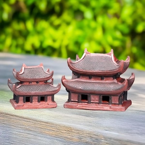 May include: Two miniature pagoda sculptures. The structures are a reddish-brown colour with dark brown roofs and intricate architectural details. The pagodas have multiple tiers and curved eaves, suggesting an Asian-inspired design.