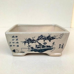 Könnte beinhalten: Rechteckiger Bonsai-Topf aus Keramik in Beige mit einer blau-weißen Illustration eines Baumes und eines Fischers. Der Topf hat eine quadratische Form und wird von vier kleinen Füßen getragen. Japanischer Text ist auf den Seiten sichtbar.