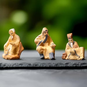 May include: Three small, ceramic figurines of seated Asian figures. Each figure is dressed in a robe with details in black and blue. The figures have long beards and are in various poses. The figurines are on a dark gray surface with a blurred green background.