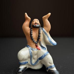 May include: A ceramic figurine of a man in a meditative pose, with arms raised. The figure has a beard and is wearing a white robe with blue trim and black shoes. A black beaded necklace hangs around the neck. The figurine is on a black surface.