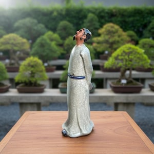 May include: A ceramic figurine of a person standing with their head tilted upward, wearing a long, light-colored robe with a fine crackle glaze finish and a dark blue belt. The figure has dark hair and is positioned on a flat wooden surface.
