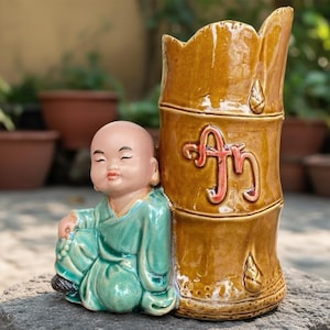 Könnte beinhalten: Eine Keramikfigur eines sitzenden Mönchs in einem tealfarbenen Gewand neben einer braunen Vase im Bambus-Stil. Die Vase hat rote und goldene Akzente. Der Mönch ist kahlköpfig mit einem friedlichen Ausdruck. Die Vase hat eine einzigartige Form und Design.