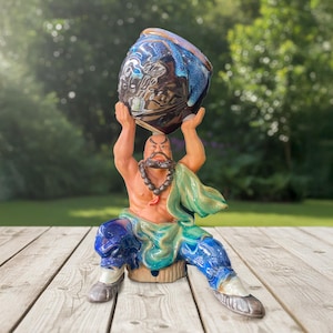 May include: A ceramic figurine of a bearded figure in a seated pose, wearing blue trousers and a green robe, holding a dark blue and brown glazed bowl above their head. The figure features a dark beaded necklace and is painted in vibrant colours.